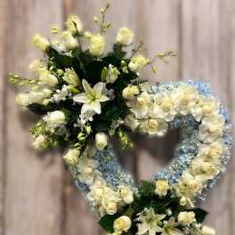 Heart-shaped wreath of white roses, lilies, and light blue hydrangeas on a stand