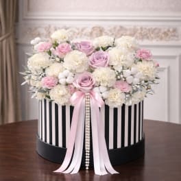 Round hat box of white carnations with pink and lavender roses, trimmed with pink ribbons and pearls