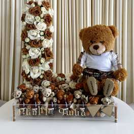 Tall number-shaped arrangement of white and caramel roses with teddy bears and mini bottles on a clear tray.