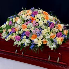 Large pastel casket spray of roses and mixed flowers in peach, purple, white, and blue on a wood casket