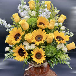 Arrangement of yellow sunflowers and roses with white blooms in a rustic clay pot on a wood slab base