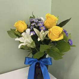 Arrangement of yellow roses, white lilies, and purple asters in a dark vase with a blue ribbon bow