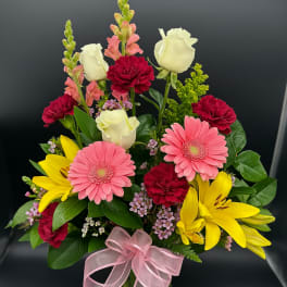 Mixed bouquet with pink gerbera daisies, yellow lilies, white roses, and red carnations in a vase with a pink bow.