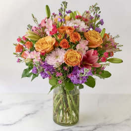 Bright mixed bouquet of orange roses, pink lilies, and purple blooms in a clear glass vase