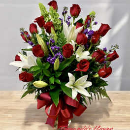 Tall arrangement of red roses, white lilies, purple flowers and green bells in a glass vase with red bow