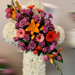 Standing white floral cross with cluster of purple roses, orange lilies, and pink gerberas.