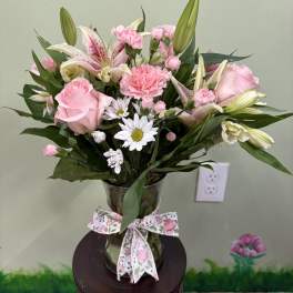 Pink roses, lilies, carnations, and white daisies in a clear glass vase with a floral bow.