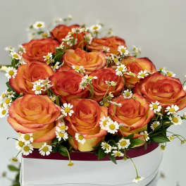 Low arrangement of orange and yellow roses with white daisies in a white hat box container