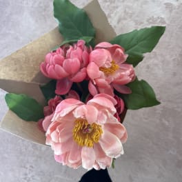 Small bouquet of pink peonies with yellow centers wrapped in kraft paper, held against a light background