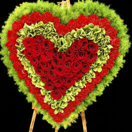 Heart-shaped standing floral spray of red roses with green and red flower borders on a wooden easel.