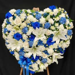 Heart-shaped blue and white standing floral wreath with roses, lilies, and carnations on a wooden easel.