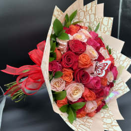 Hand-tied bouquet of red, pink, and peach roses wrapped in patterned paper with a red bow and butterfly accents