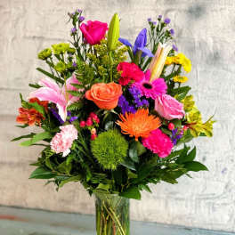 Tall mixed bouquet of bright pink, orange, yellow, and purple flowers in a clear glass vase.