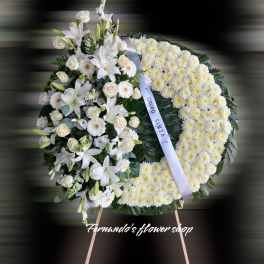 Standing white funeral wreath with lilies, roses, and chrysanthemums on an easel with a white ribbon