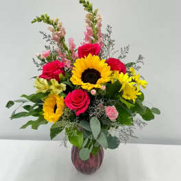 Mixed bouquet with sunflowers, hot pink roses, snapdragons, and daisies in a red glass vase