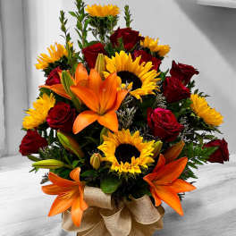 Mixed arrangement of sunflowers, orange lilies, and red roses in a glass vase with a burlap bow