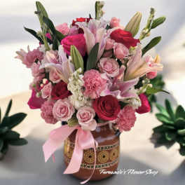 Pink and red bouquet with lilies, roses, and carnations in a painted ceramic vase with a pink bow.