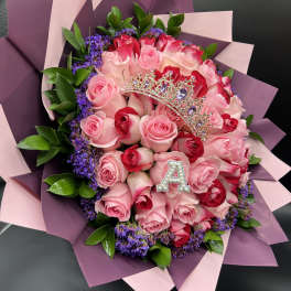 Bouquet of pink and red roses with purple filler flowers, wrapped in pink paper with a jeweled tiara and letter A.