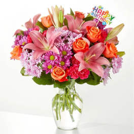 Mixed bouquet of pink lilies, roses, and daisies in a glass vase with a birthday topper