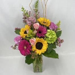 Tall mixed arrangement with sunflowers, pink gerberas, roses, and green hydrangeas in a clear glass vase