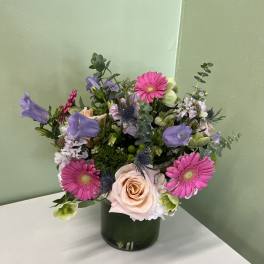 Mixed arrangement of pink gerbera daisies, peach roses, and purple blooms in a green glass cylinder vase