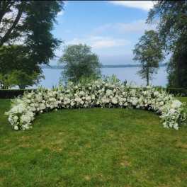 Low crescent of lush white wedding flowers arranged on a lawn overlooking a lake.