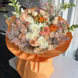 Hand-tied bouquet of peach and white roses with orange ranunculus wrapped in bright orange paper.