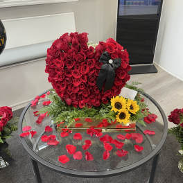 Heart-shaped red rose arrangement with a black ribbon and sunflowers on a glass table