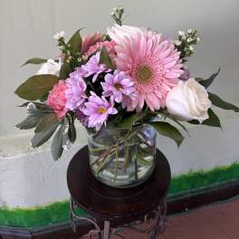 Pink gerbera daisies, roses, and lavender mums in a clear glass vase on a small stand