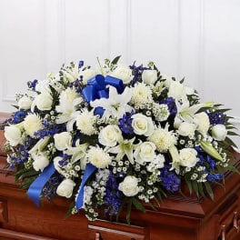 White and blue casket spray with roses, lilies, and ribbon on a polished wooden casket