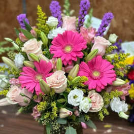 Vase arrangement with hot pink gerbera daisies, pale roses, lilies, and mixed blooms in a green textured glass vase