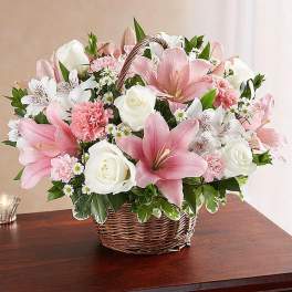 Pink lilies, white roses, and mixed blooms arranged in a wicker basket on a wooden table