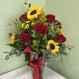 Tall vase of red roses and yellow sunflowers with small yellow daisies and a red bow.