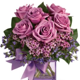Lavender roses with small pink and purple blooms in a lavender glass cube vase with ribbon.