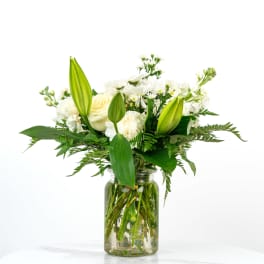 White lilies, roses, and other white flowers arranged in a clear glass jar vase