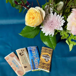Floral arrangement with pink and yellow blooms beside four wrapped chocolate bars on teal fabric.