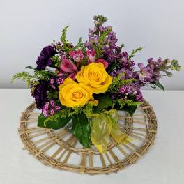 Compact arrangement of yellow roses and purple mixed flowers in a glass vase with a yellow bow