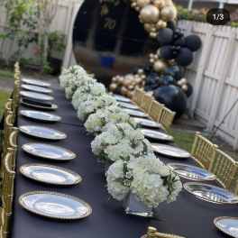 White floral centerpieces on a black table with gold plates and chairs