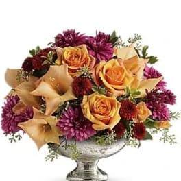 Low centerpiece of orange roses, peach calla lilies, and purple mums in a silver pedestal bowl
