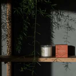 Candle in a glass jar beside its orange box on a wooden shelf