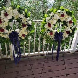 Two floral wreaths on stands with white flowers, red roses, and navy ribbons
