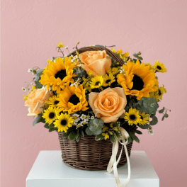 Basket arrangement of yellow sunflowers, peach roses, and small yellow daisies with a ribbon bow.