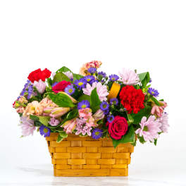 Low basket arrangement of pink daisies, red roses, red carnations, and purple asters in a woven wood basket