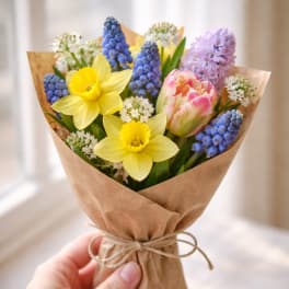 Small hand-tied bouquet of yellow daffodils, pink tulip, and blue hyacinths wrapped in brown paper