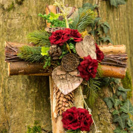 Rustic wooden cross decorated with red flowers, evergreens, bark hearts, and pinecones.