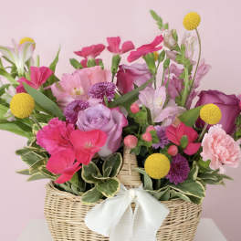 Wicker basket of pink, purple, and yellow mixed flowers with a white bow.