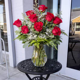 Arrangement of long-stem red roses with white filler flowers in a clear glass vase on a black metal table.