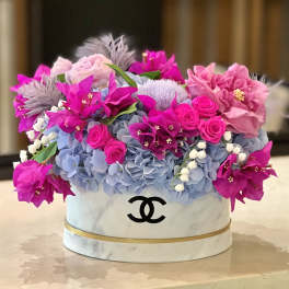 Pink and blue floral arrangement in a white round box with a black logo