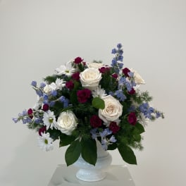 Arrangement of white roses, white daisies, burgundy carnations, and blue delphinium in a white pedestal vase