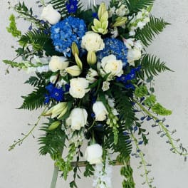 Tall floral standing spray with white roses, blue hydrangeas, and a silver cross